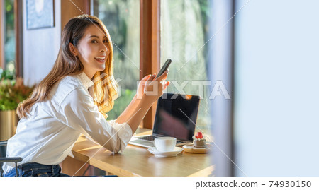 Portrait Young Asian woman holding and drinking a cup of coffee and working with technology laptop at a coffee shop. freelancer and entrepreneur working by connecting to internet via computer 74930150