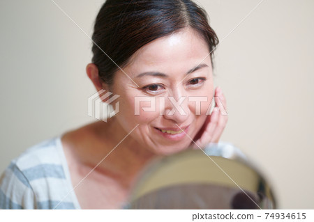看鏡子和把她的手放在她的臉頰上的一名微笑的中年婦女 看鏡子和把她的手放在她的臉頰上的一名微笑的中年婦女 74934615