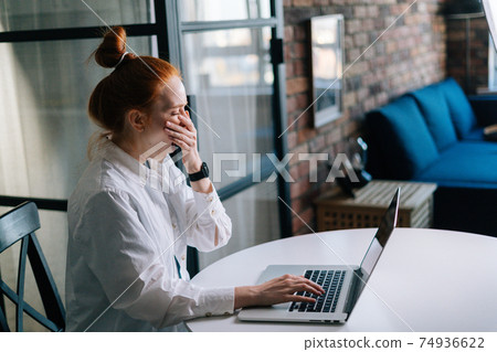 Side view of sleepy redhead young woman yawning while working on laptop computer sitting at table Side view of sleepy redhead young woman yawning while working on laptop computer sitting at table 74936622