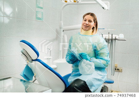a female dentist with a medical mask and rubber gloves in her hands sits in her office 74937468