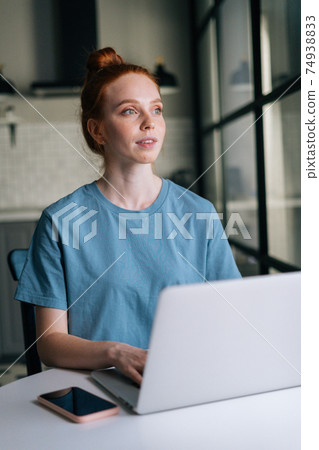 Portrait of Pensive redhead young woman working typing on laptop computer sitting at table 74938833