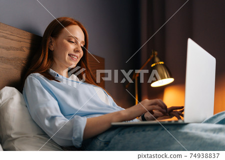 Close-up of relaxed redhead young woman looking on laptop screen while lying on bed. 74938837