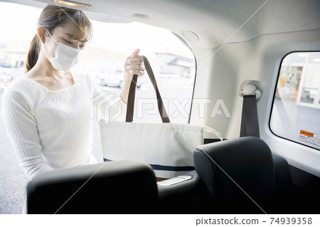A woman in a mask who puts her shopping baggage in the trunk of a car 74939358