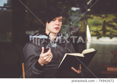 Young black haired man witn piercing reeding a book and smoking tabacco pipe in outdoor cafe. Young black haired man witn piercing reeding a book and smoking tabacco pipe in outdoor cafe. 74942672