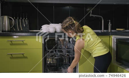 Smart girl learning to use dishwasher. Stylish modern Built In Kitchen Appliances in green black. Child is putting dirty dishes. Smart girl learning to use dishwasher. Stylish modern Built In Kitchen Appliances in green black. Child is putting dirty dishes. 74946426