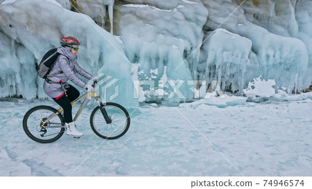 Woman is riding bicycle near the ice grotto. The rock with ice c Woman is riding bicycle near the ice grotto. The rock with ice c 74946574