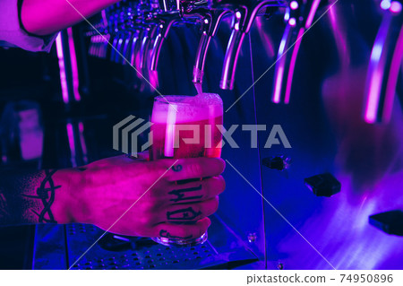 Hands of barman pouring lager beer from tap to glass in neon light. Hands of barman pouring lager beer from tap to glass in neon light. 74950896
