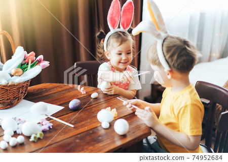 A boy and a girl are painting Easter eggs for the holiday at a wooden table. Children have bunny ears on their heads 74952684