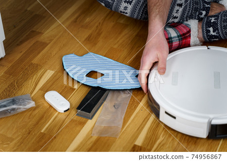 Caucasian young man in christmas pajamas doing vacuum cleaner robot maintenance. Male in winter patterned sleepwear and red socks slippers starts automatic vacuum cleaner. Robot vacuum cleaner repair 74956867