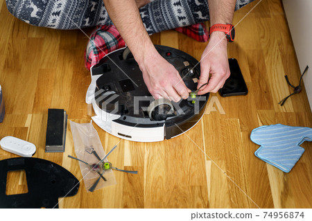 A young father in sleepwear with a winter ornament and Christmas funny socks serves a roobot vacuum cleaner for cleaning the house for the New Year and Christmas holidays. New tech for households 74956874