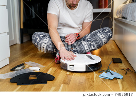 A young father in sleepwear with a winter ornament and Christmas funny socks serves a roobot vacuum cleaner for cleaning the house for the New Year and Christmas holidays. New tech for households 74956917