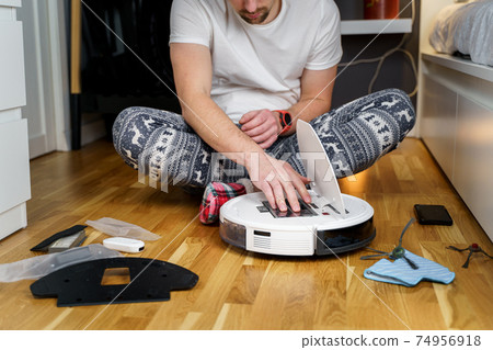 Young caucasian man doing cleaning for the holiday christmas in winter. A male in funny home clothes, pajamas with winter ornament collects and sets up a robot vacuum cleaner at home on the floor Young caucasian man doing cleaning for the holiday christmas in winter. A male in funny home clothes, pajamas with winter ornament collects and sets up a robot vacuum cleaner at home on the floor 74956918