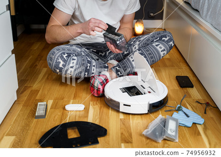 Young caucasian man doing cleaning for the holiday christmas in winter. A male in funny home clothes, pajamas with winter ornament collects and sets up a robot vacuum cleaner at home on the floor Young caucasian man doing cleaning for the holiday christmas in winter. A male in funny home clothes, pajamas with winter ornament collects and sets up a robot vacuum cleaner at home on the floor 74956932