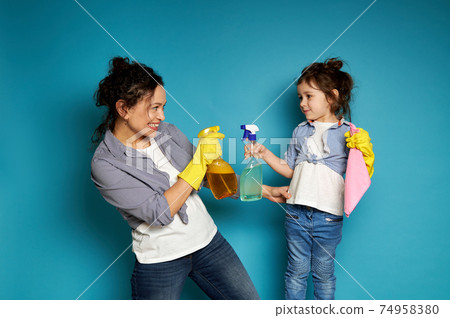 Cheerful mother and daughter hold a cleaning spray while playing with detergents. Household together. Cheerful mother and daughter hold a cleaning spray while playing with detergents. Household together. 74958380
