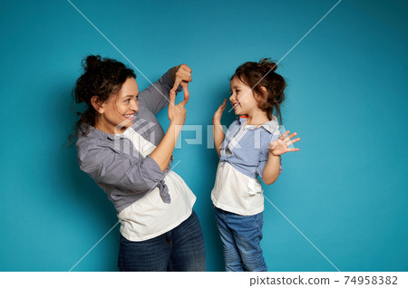Side view of a smiling curly woman looking at her cute baby girl through her fingers, framing her face. Blue background with copy space Side view of a smiling curly woman looking at her cute baby girl through her fingers, framing her face. Blue background with copy space 74958382