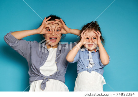 Happy woman and her daughter looking at camera through their fingers in ok gesture, imitating binoculars. Copy space, blue background. Happy woman and her daughter looking at camera through their fingers in ok gesture, imitating binoculars. Copy space, blue background. 74958384