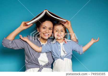 Mom and daughter happily smile at the camera while holding a book over their heads in the form of a roof from the house Mom and daughter happily smile at the camera while holding a book over their heads in the form of a roof from the house 74958385