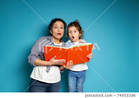 Surprised woman and child looking at camera holding an orange book in their book. Blue background with copy space. Surprised woman and child looking at camera holding an orange book in their book. Blue background with copy space. 74958386