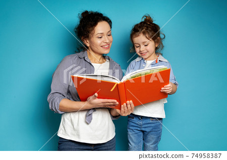 Woman and girl equally dressed hold a book in their hands, smiling tenderly, looking at it. Blue background with copy space Woman and girl equally dressed hold a book in their hands, smiling tenderly, looking at it. Blue background with copy space 74958387