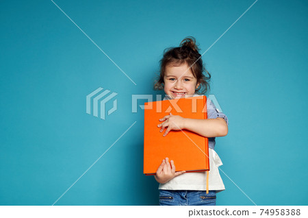 Adorable little girl gently hugs an orange book and cute smiles with toothy smile looking at camera while standing on blue background with copy space Adorable little girl gently hugs an orange book and cute smiles with toothy smile looking at camera while standing on blue background with copy space 74958388