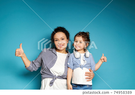 Beautiful woman and daughter equally dressed showing thumbs up and cute smiling while posing to camera on blue background with copy space Beautiful woman and daughter equally dressed showing thumbs up and cute smiling while posing to camera on blue background with copy space 74958389