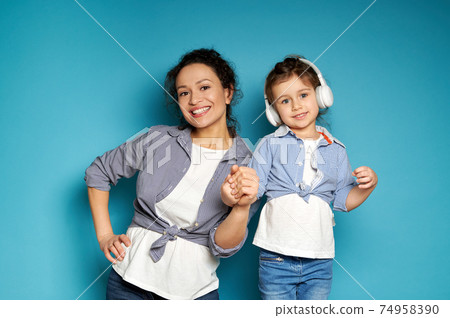 Attractive woman posing in front of the camera with a toothy smile on a blue background with her little daughter in headphones Attractive woman posing in front of the camera with a toothy smile on a blue background with her little daughter in headphones 74958390