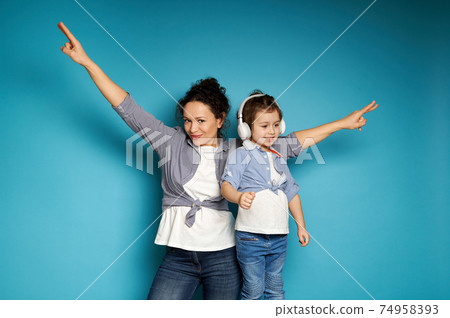Adorable mother and daughter wearing headphones, equally dressed singing and dancing against a blue background with copy space. Adorable mother and daughter wearing headphones, equally dressed singing and dancing against a blue background with copy space. 74958393