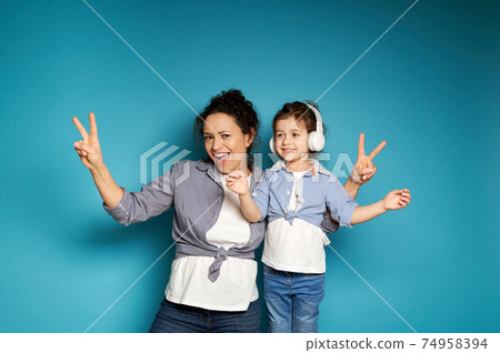 A woman and a girl wearing headphones, equally dressed singing and dancing, posing against a blue background with copy space. A woman and a girl wearing headphones, equally dressed singing and dancing, posing against a blue background with copy space. 74958394