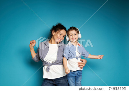 Adorable baby girl with headphones dancing near her mother hugging her on a blue background with copy space Adorable baby girl with headphones dancing near her mother hugging her on a blue background with copy space 74958395