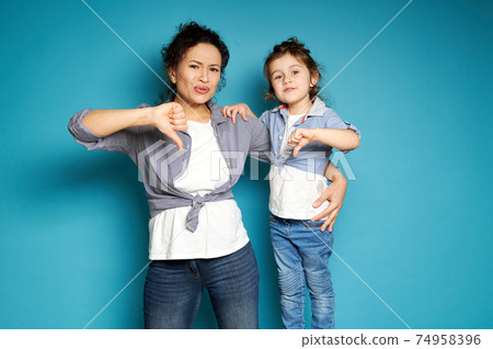Beautiful mom and daughter equally dressed showing thumbs down and expressing dissatisfaction while posing to camera on blue background with copy space Beautiful mom and daughter equally dressed showing thumbs down and expressing dissatisfaction while posing to camera on blue background with copy space 74958396