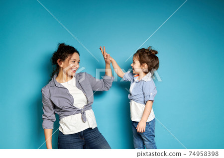 Young curly woman and cute little girl look at each other and clapping hands gesturing "high five". Mother and and daughter on blue background Young curly woman and cute little girl look at each other and clapping hands gesturing "high five". Mother and and daughter on blue background 74958398