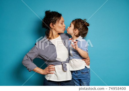 Curly brunette woman and her cute baby girl kissing while looking to each other on blue background Curly brunette woman and her cute baby girl kissing while looking to each other on blue background 74958400