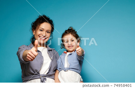 Beautiful mom and daughter equally dressed showing thumbs up and cute smiling while posing to camera on blue background with copy space Beautiful mom and daughter equally dressed showing thumbs up and cute smiling while posing to camera on blue background with copy space 74958401