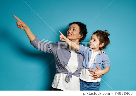 Young smiling woman and her cute little daughter pointing with forefinger on blue background Young smiling woman and her cute little daughter pointing with forefinger on blue background 74958403