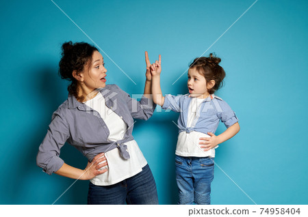 Young woman and her baby girl alike dressed looking at each other and pointing their index finger up. Blue background with copy space Young woman and her baby girl alike dressed looking at each other and pointing their index finger up. Blue background with copy space 74958404
