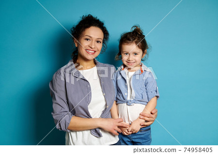 Portrait of a happy smiling woman hugging her daughter standing on blue background with copy space. Love, care and tenderness concept Portrait of a happy smiling woman hugging her daughter standing on blue background with copy space. Love, care and tenderness concept 74958405
