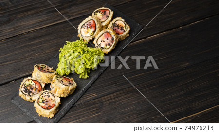 Custom sushi roll in tempura with nori, fresh salmon, tuna, avocado, masago caviar, drizzled with pineapple sauce with salad pouring as decoration on a black plate on a wooden table and background. Custom sushi roll in tempura with nori, fresh salmon, tuna, avocado, masago caviar, drizzled with pineapple sauce with salad pouring as decoration on a black plate on a wooden table and background. 74976213
