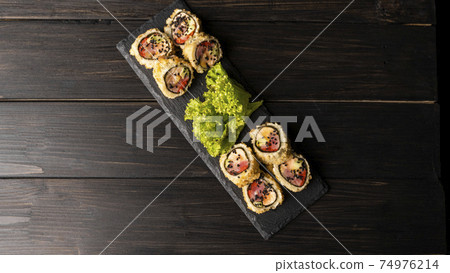 Custom sushi roll in tempura with nori, fresh salmon, tuna, avocado, masago caviar, drizzled with pineapple sauce with salad pouring as decoration on a black plate on a wooden table and background. Custom sushi roll in tempura with nori, fresh salmon, tuna, avocado, masago caviar, drizzled with pineapple sauce with salad pouring as decoration on a black plate on a wooden table and background. 74976214