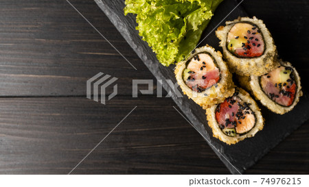 Custom sushi roll in tempura with nori, fresh salmon, tuna, avocado, masago caviar, drizzled with pineapple sauce with salad pouring as decoration on a black plate on a wooden table and background. 74976215