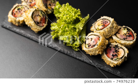 Custom sushi roll in tempura with nori, fresh salmon, tuna, avocado, masago caviar, drizzled with pineapple sauce with salad pouring as decoration on a black plate on a black table and background. Custom sushi roll in tempura with nori, fresh salmon, tuna, avocado, masago caviar, drizzled with pineapple sauce with salad pouring as decoration on a black plate on a black table and background. 74976219