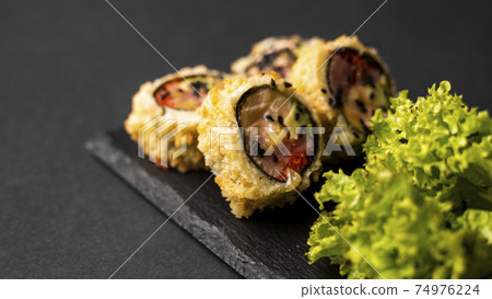 Custom sushi roll in tempura with nori, fresh salmon, tuna, avocado, masago caviar, drizzled with pineapple sauce with salad pouring as decoration on a black plate on a black table and background. 74976224