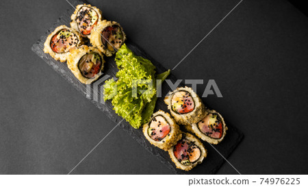 Custom sushi roll in tempura with nori, fresh salmon, tuna, avocado, masago caviar, drizzled with pineapple sauce with salad pouring as decoration on a black plate on a black table and background. Custom sushi roll in tempura with nori, fresh salmon, tuna, avocado, masago caviar, drizzled with pineapple sauce with salad pouring as decoration on a black plate on a black table and background. 74976225