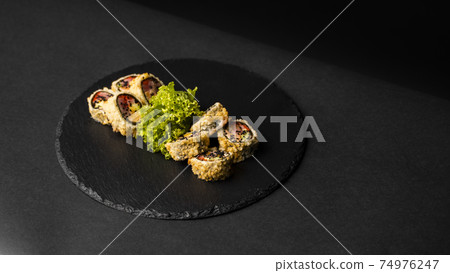 Custom sushi roll in tempura with nori, fresh salmon, tuna, avocado, masago caviar, drizzled with pineapple sauce with salad pouring as decoration on a black plate on a black table and background. Custom sushi roll in tempura with nori, fresh salmon, tuna, avocado, masago caviar, drizzled with pineapple sauce with salad pouring as decoration on a black plate on a black table and background. 74976247