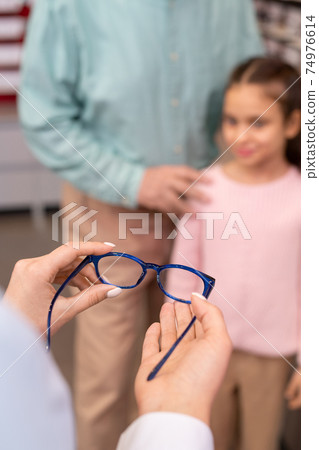 Hands of young female optician offering pair of new eyeglasses to little girl 74976614