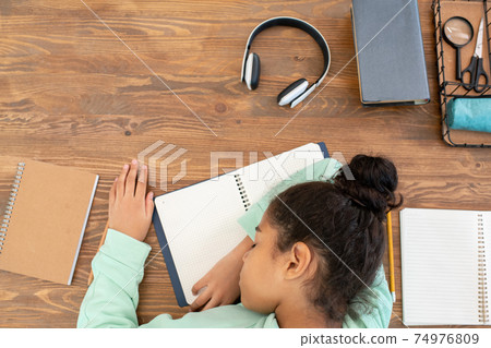 Overview of tired mixed-race schoolgirl keeping her head on open copybook Overview of tired mixed-race schoolgirl keeping her head on open copybook 74976809