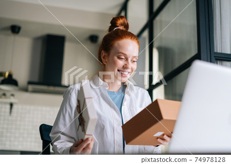 Low-angle shot of cheerful redhead young woman opening gift box with present during video call 74978128