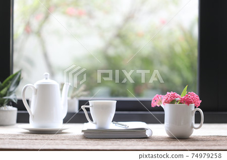 White Tea cup and teapot with pink Panama Rose and notebook in front of winwow glass White Tea cup and teapot with pink Panama Rose and notebook in front of winwow glass 74979258