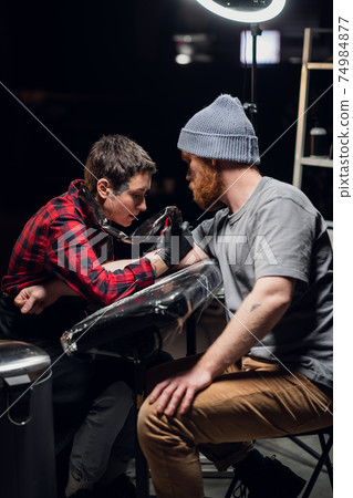 In the interior of a tattoo parlor, a young man with a beard is tattooed on his arm using the stick and poke method. Young artist girl in a checkered shirt. 74984877