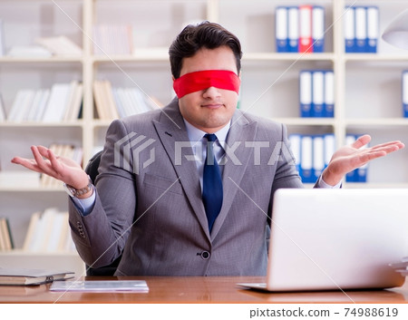 Blindfold businessman sitting at desk in office Blindfold businessman sitting at desk in office 74988619