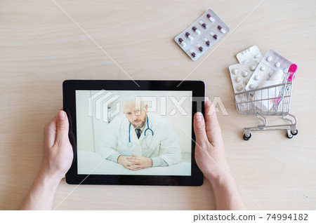 The patient communicates with a male therapist via video chat on a digital tablet. Woman at online consultation with a doctor. Online pharmacy concept. Mini medicine trolley. 74994182